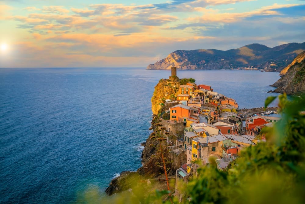 A high-angle view of Vernazza, a colorful cliffside village in Cinque Terre, Italy, with historic buildings and a stone tower overlooking the deep blue sea at sunrise or sunset.
