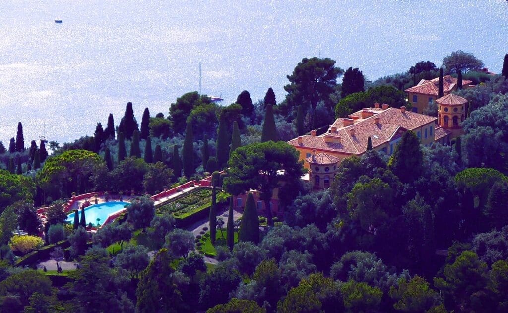An aerial view of Villa Leopolda, a grand estate with red-tiled roofs and a large swimming pool, nestled amidst lush trees overlooking the Mediterranean Sea.