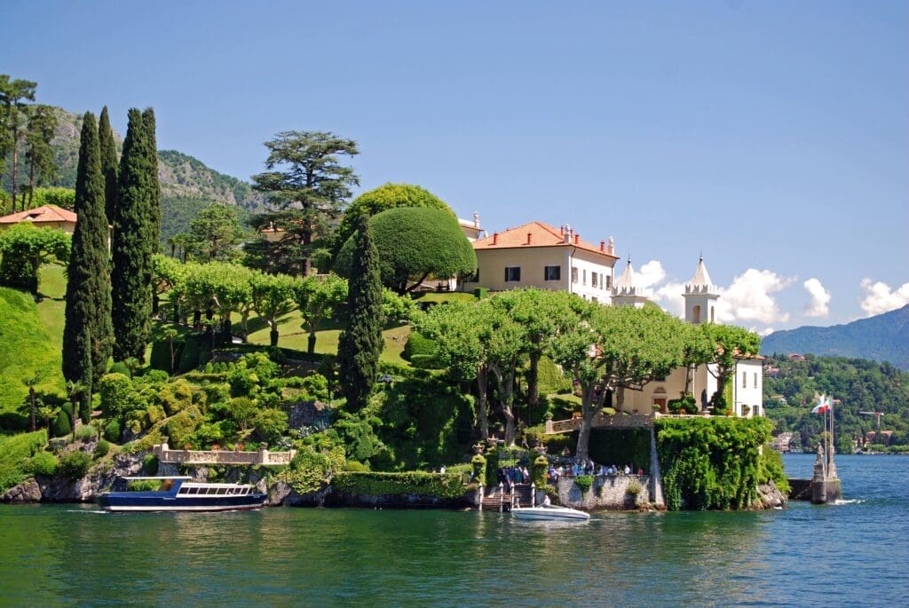 A panoramic view of Villa del Balbianello on Lake Como, Italy, featuring a grand white villa nestled in lush terraced gardens with cypress trees, overlooking the calm lake with docked boats.
