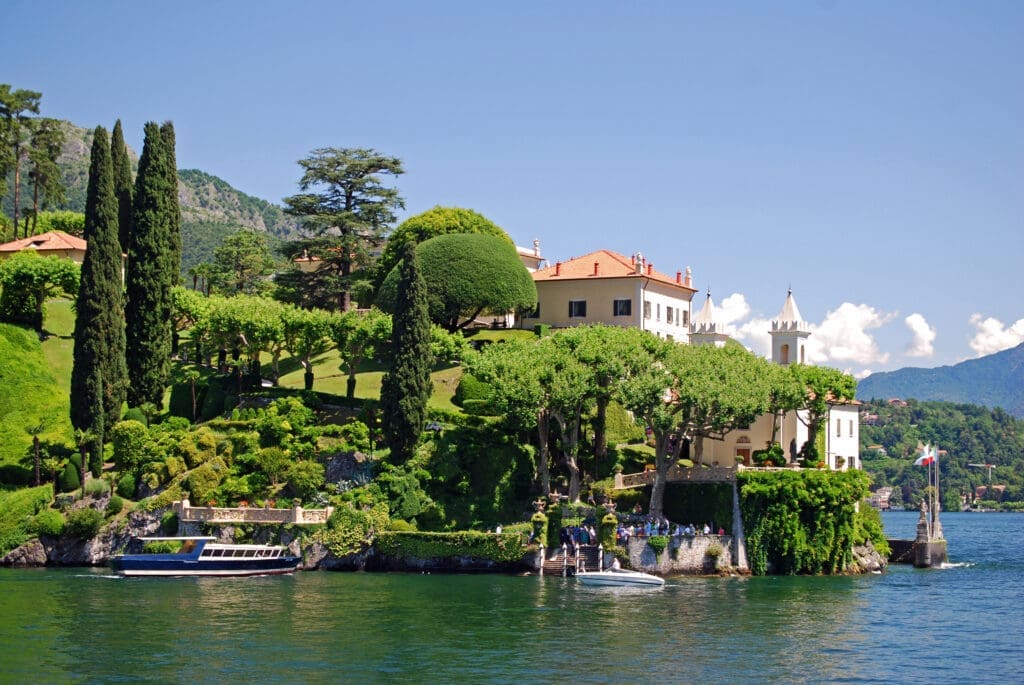 A panoramic view of Villa del Balbianello on Lake Como, Italy, featuring a grand white villa nestled in lush terraced gardens with cypress trees, overlooking the calm lake with docked boats.