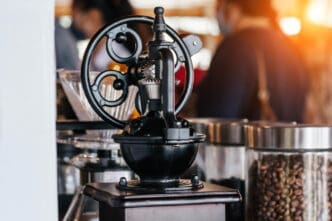 A vintage black coffee grinder with a hand crank and a wheel is positioned on a wooden base, with a glass jar of coffee beans and blurred people in the background, in a sunlit cafe.