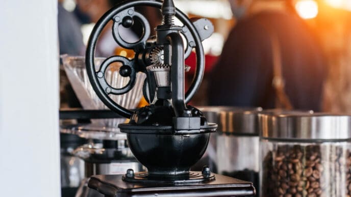 A vintage black coffee grinder with a hand crank and a wheel is positioned on a wooden base, with a glass jar of coffee beans and blurred people in the background, in a sunlit cafe.