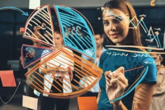 A young woman in a blue t-shirt draws a large pie chart and other diagrams on a transparent glass board with markers, smiling at the camera, with blurred colleagues in the background.