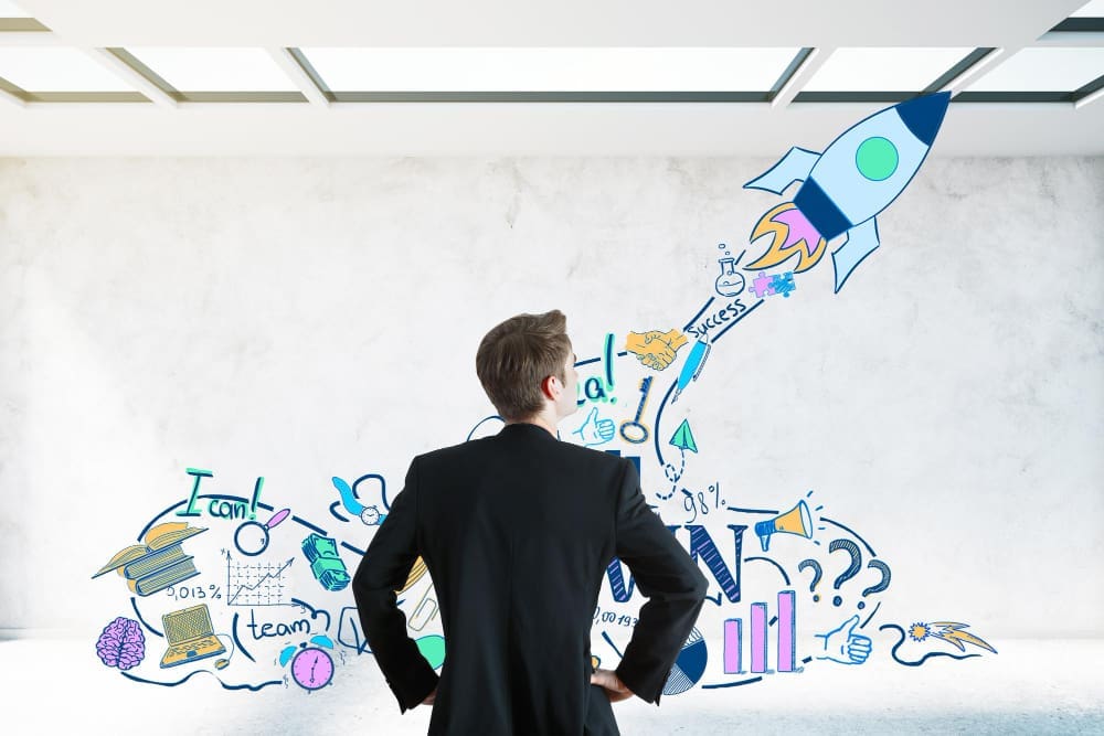 A businessman seen from behind, looking at a wall covered with colorful doodles of a launching rocket, ideas, and business concepts, symbolizing goal setting.