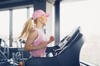 A young woman with long blonde hair and a pink baseball cap runs on a treadmill in a gym, wearing a pink tank top and earphones, looking focused as she runs next to a large window.