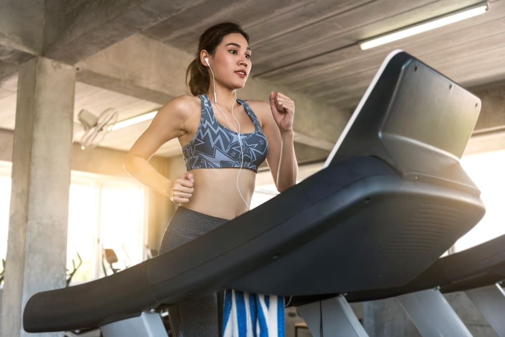 A young woman who appears to be of East Asian descent is running on a treadmill in a gym, wearing a patterned sports bra and earphones, with a focused expression.