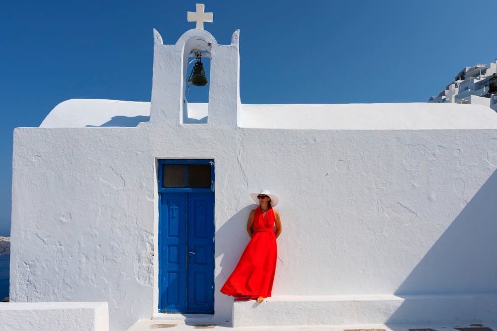 A woman in a vibrant red dress and white hat stands leaning against a white church with a blue door and a bell tower, typical of Santorini, Greece, under a clear blue sky.