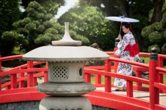 A woman in a white floral kimono with a red obi stands on a red Japanese bridge, holding a white parasol, in a traditional garden setting with a stone lantern in the foreground.