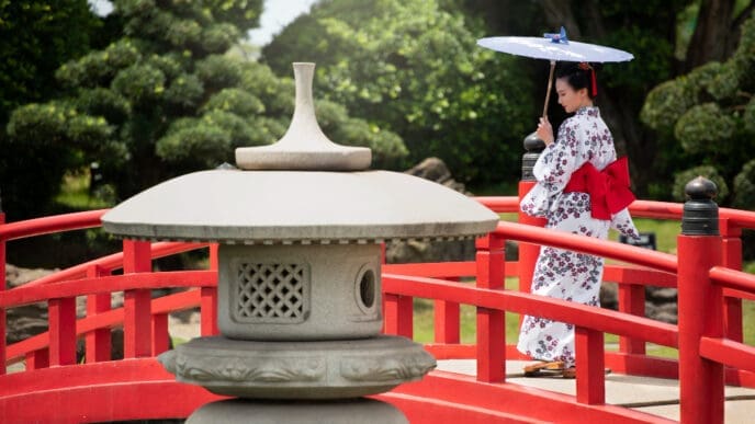 A woman in a white floral kimono with a red obi stands on a red Japanese bridge, holding a white parasol, in a traditional garden setting with a stone lantern in the foreground.