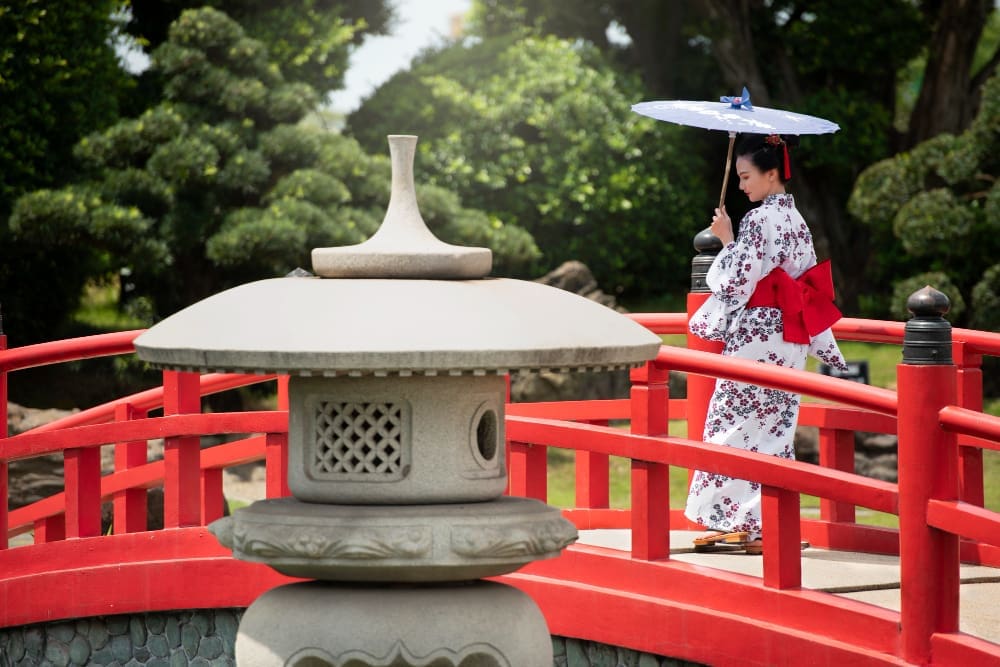 A woman in a white floral kimono with a red obi stands on a red Japanese bridge, holding a white parasol, in a traditional garden setting with a stone lantern in the foreground.