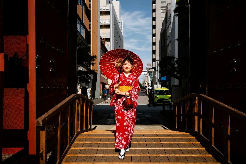 A woman wearing a vibrant red floral kimono and holding a red wagasa umbrella walks on a traditional wooden bridge in Kyoto's Gion district, with modern city buildings in the background.