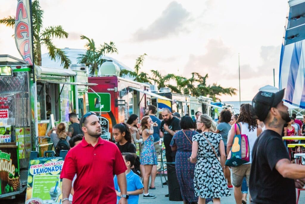 A bustling scene at a food truck festival in Wynwood, Miami, with a crowd of people walking past a line of food trucks, with palm trees and a cloudy sky at sunset in the background.