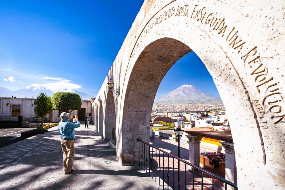 A man taking a photo through the stone arches of Yanahuara viewpoint, with the city of Arequipa and the Misti volcano in the background.