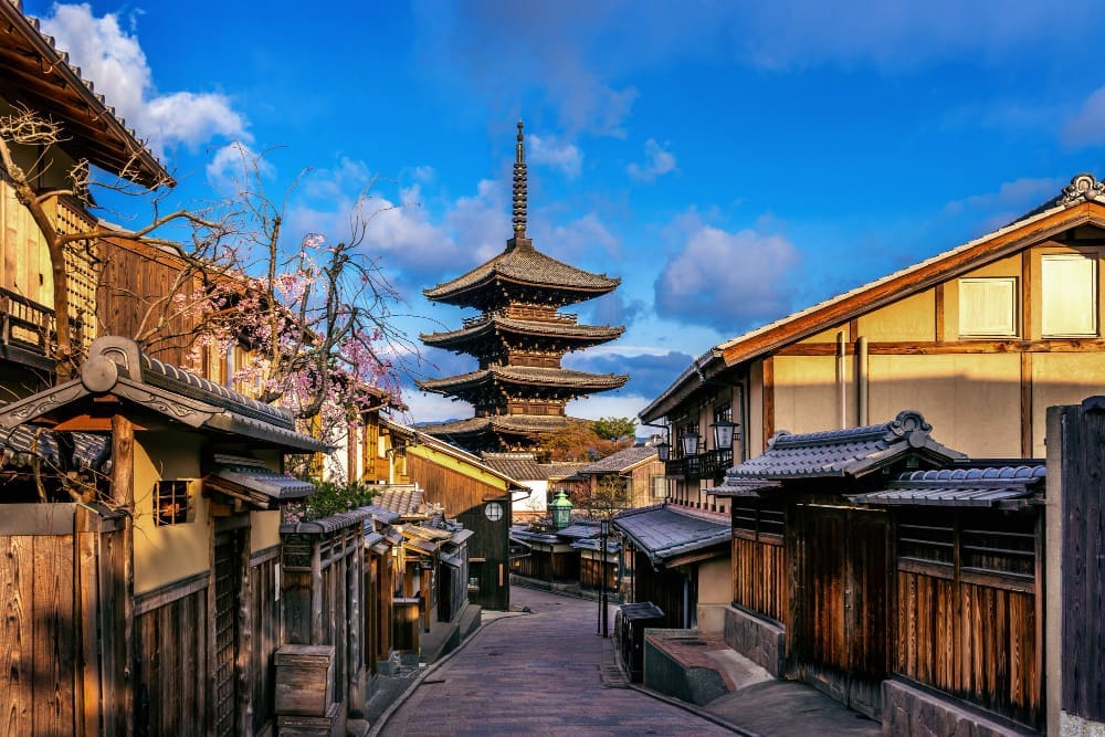 A traditional Japanese street (Sannenzaka) in Kyoto, lined with historic wooden buildings and blooming cherry blossoms, leading towards the iconic Yasaka Pagoda under a blue sky.