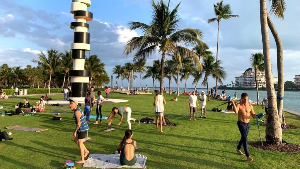People practicing yoga, slacklining, and relaxing on the grass at South Pointe Park, Miami Beach, near a black and white striped sculpture, with palm trees and water in the background.
