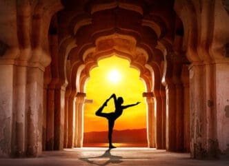 A silhouette of a person performing a yoga pose (Natarajasana or dancer pose) in the center of a long archway of ancient stone columns, with a bright orange sunset over water and distant mountains in the background.