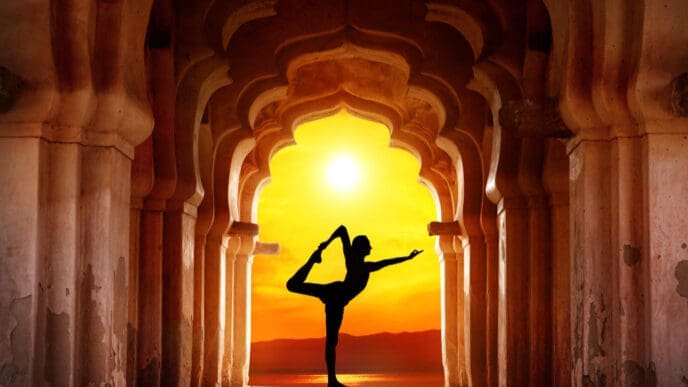 A silhouette of a person performing a yoga pose (Natarajasana or dancer pose) in the center of a long archway of ancient stone columns, with a bright orange sunset over water and distant mountains in the background.