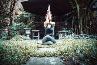 A person performs a yoga pose (Gomukhasana with Garudasana arms) in a lush outdoor garden, seated on the ground with their arms raised and intertwined above their head, under a large black umbrella.