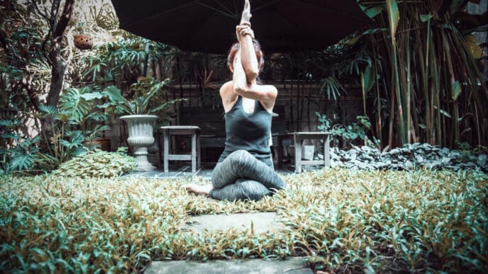 A person performs a yoga pose (Gomukhasana with Garudasana arms) in a lush outdoor garden, seated on the ground with their arms raised and intertwined above their head, under a large black umbrella.