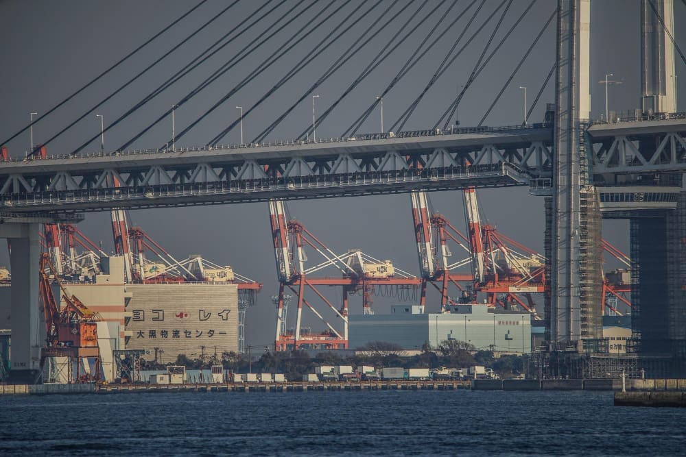 The impressive Yokohama Bay Bridge spans across the frame, with large red and white gantry cranes and industrial buildings of a port facility visible beneath it, all set against a dark sky and calm water.