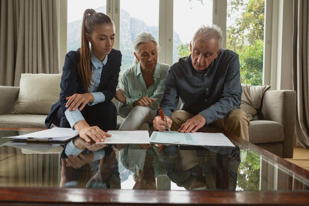 An elderly couple and a real estate agent review documents together in a living room.