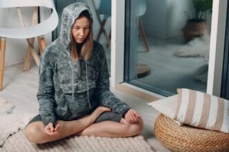 An adult woman practices the lotus yoga pose in her living room, following online tutorials on a laptop.