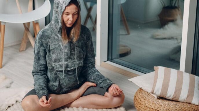 An adult woman practices the lotus yoga pose in her living room, following online tutorials on a laptop.