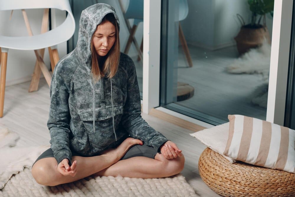 An adult woman practices the lotus yoga pose in her living room, following online tutorials on a laptop.