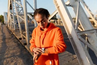 An African American woman in athletic wear listens to music on earbuds and monitors her workout on a smartwatch while exercising on a weathered bridge.