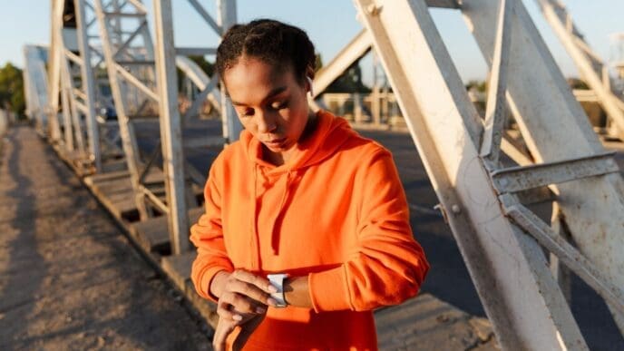 An African American woman in athletic wear listens to music on earbuds and monitors her workout on a smartwatch while exercising on a weathered bridge.