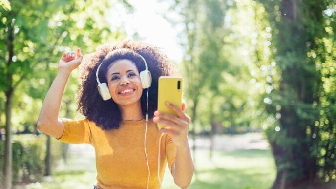 A Black girl with an afro dances joyfully in a park, wearing headphones and listening to music.
