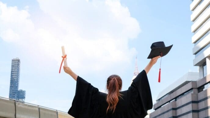 Two Asian women in graduation gowns hold their diplomas and smile.