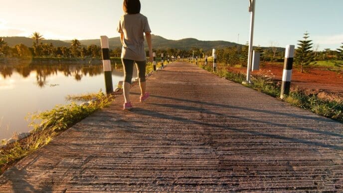 A young Asian woman in fitness attire walks along a path in a tropical park after running.