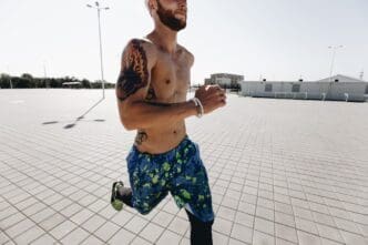 Athletic man with tattoos and a headband runs on paving slabs, wearing black leggings and blue shorts on a sunny day.