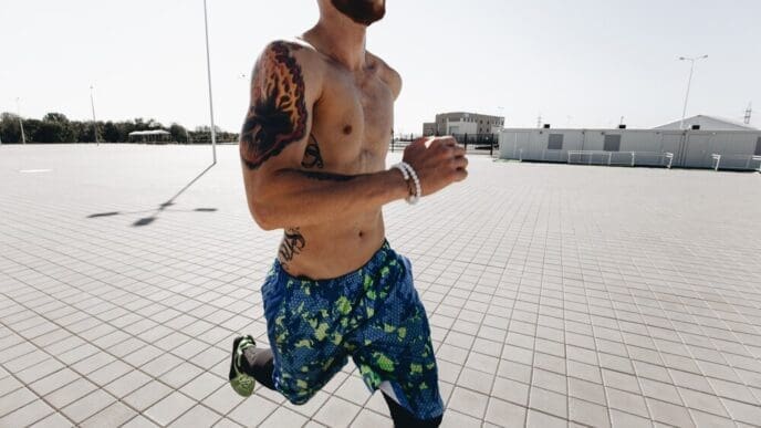 Athletic man with tattoos and a headband runs on paving slabs, wearing black leggings and blue shorts on a sunny day.