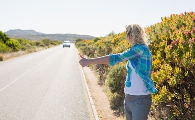Blonde woman with long hair and sunglasses stands on a rural road, holding her thumb out to hitchhike.