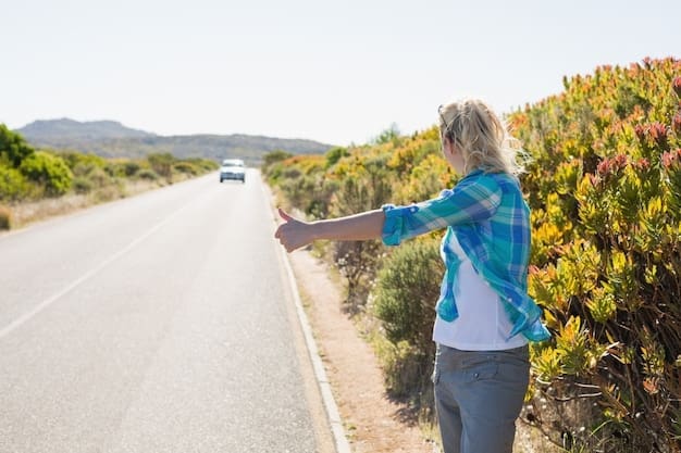 Blonde woman with long hair and sunglasses stands on a rural road, holding her thumb out to hitchhike.