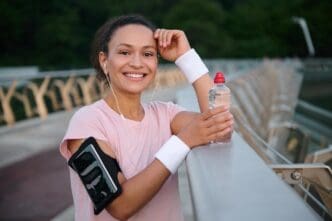 A smiling young sportswoman with a smartphone on her arm rests on a city bridge after a morning run, looking at the camera.