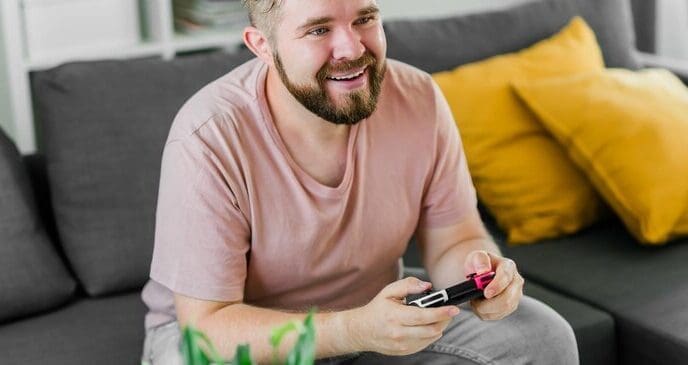 A young man smiles while playing video games on a sofa, with copy space suggesting themes of gaming addiction.