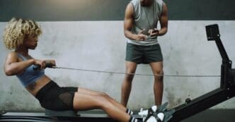A young woman exercises on a cable row machine in a gym, with a fitness trainer observing.