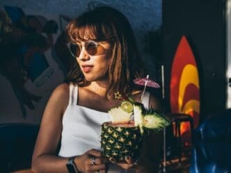 A smiling Asian woman in sunglasses and a white sleeveless top holds a pineapple juice cocktail in the summer sun.
