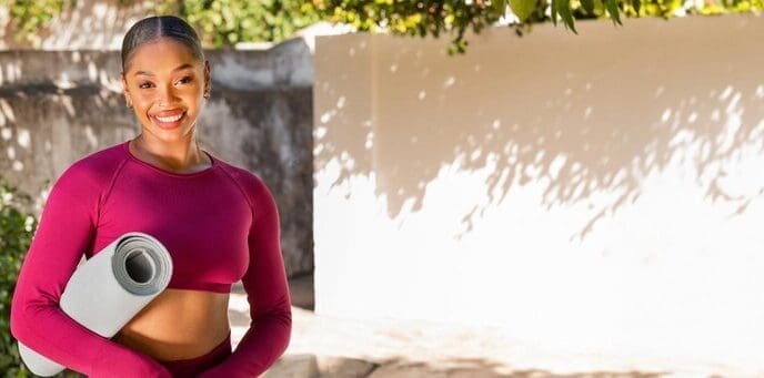 A young Black woman in pink workout clothes smiles while exercising outdoors in a garden.