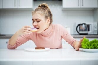 A young woman stands between healthy and junk food, appearing to contemplate her choices.