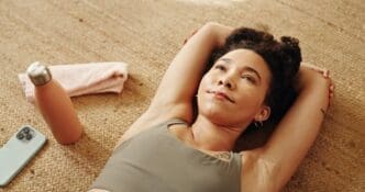 A woman in workout attire contemplates while resting on a gym floor, a water bottle nearby.