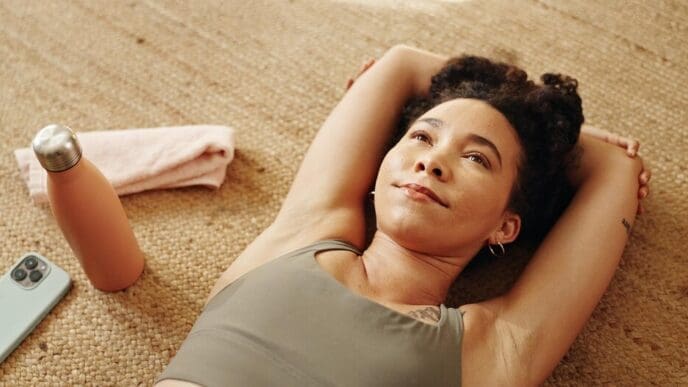 A woman in workout attire contemplates while resting on a gym floor, a water bottle nearby.