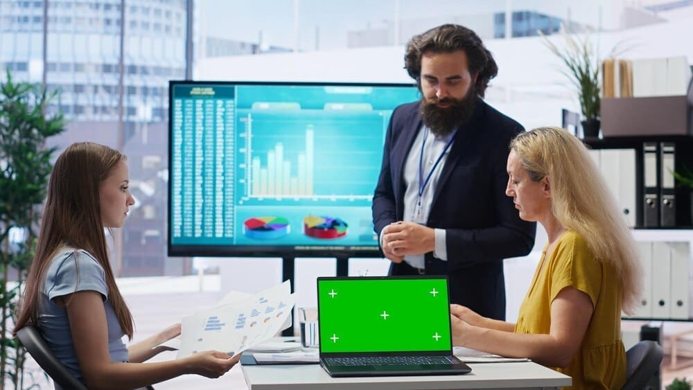 A broker points to stock exchange charts displayed on a laptop with a green screen background.