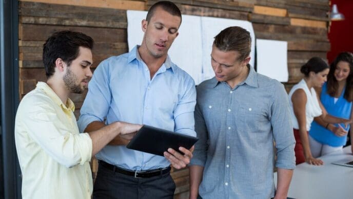 Business executives gather around a table, looking at and discussing content on a digital tablet.