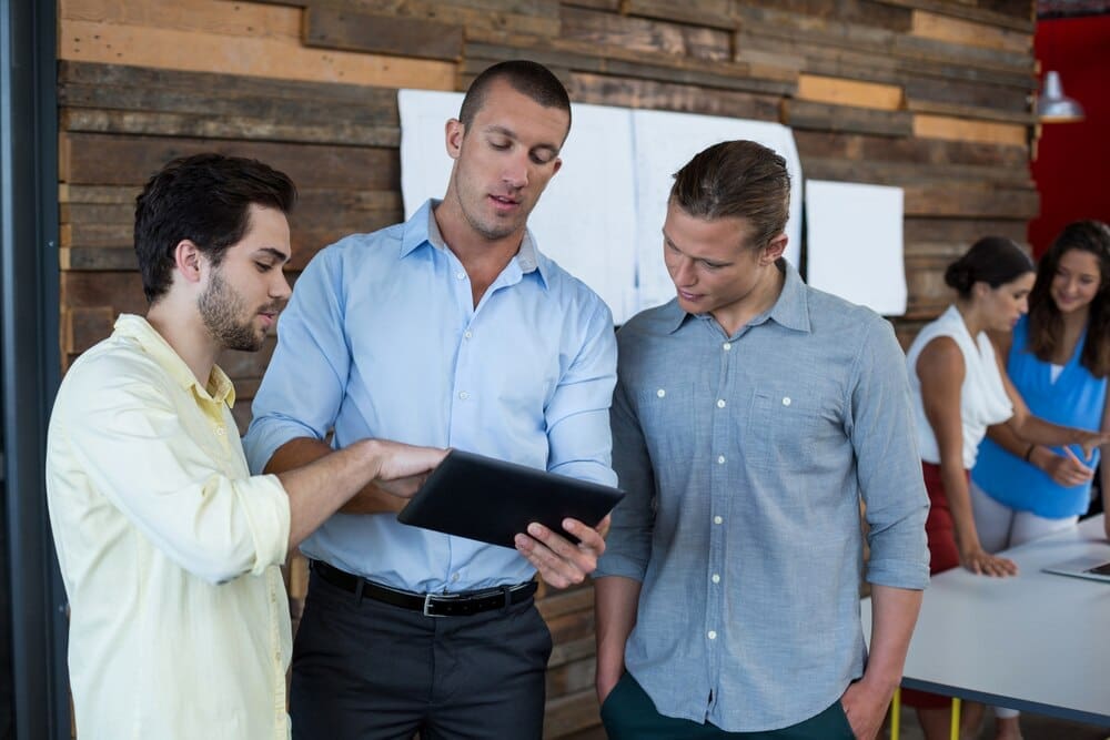Business executives gather around a table, looking at and discussing content on a digital tablet.