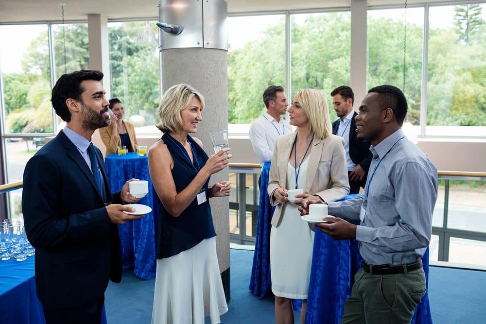 Business executives in suits and ties converse and gesture while sitting around a table with coffee cups.