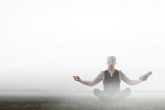 A businessman in a suit sits cross-legged on a yoga mat, meditating in the lotus position.